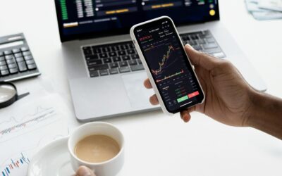 A person trades stocks on a smartphone with a laptop, coffee cup, calculator, and documents in view.